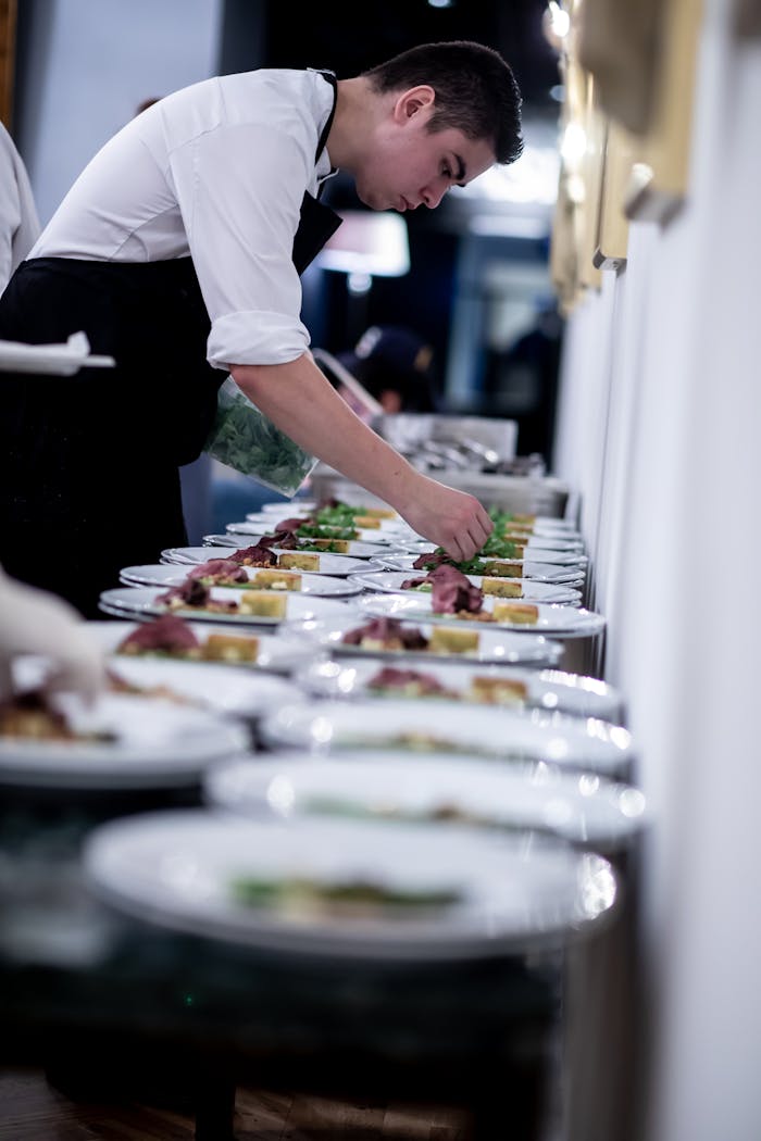 Side view of male cook in apron serving many plates with delicious food while preparing lunch in restaurant at big table
