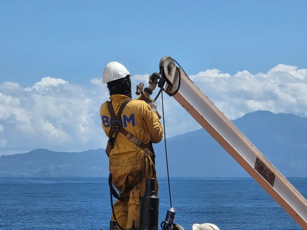 Training & Courses A worker in safety gear secures equipment on a boat against a scenic ocean backdrop in Mozambique.