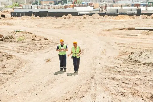 Site Supervision & Management Professional Programme-(FULL DETAILS) Two workers in safety gear walking across a construction site under a clear sky.